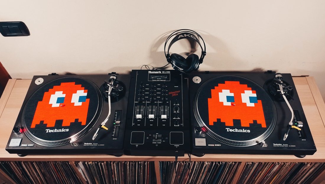 Due giradischi Technics SL-1210 MKII con tappetini a tema Pac-Man, affiancati a un mixer Numark e a una cuffia, sopra uno scaffale pieno di vinili.